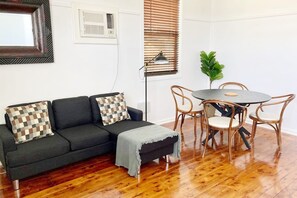 Uma sala de estar com um sofá cinza escuro, almofadas xadrez, uma mesa pequena com cadeiras de vime, uma planta em vaso e um abajur.