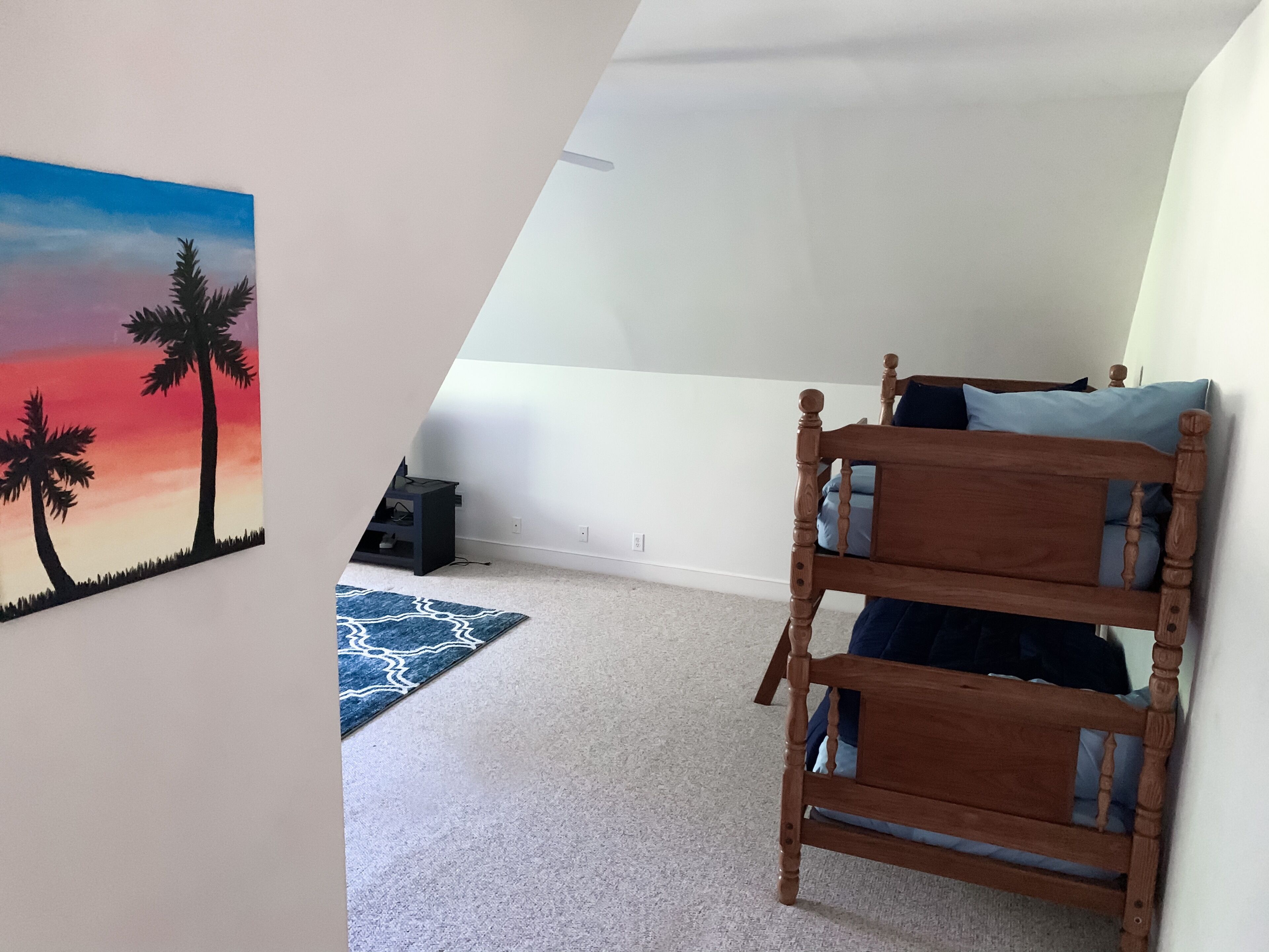 A bedroom with a bunk bed, a painting of palm trees, a blue rug, and a desk with a chair.