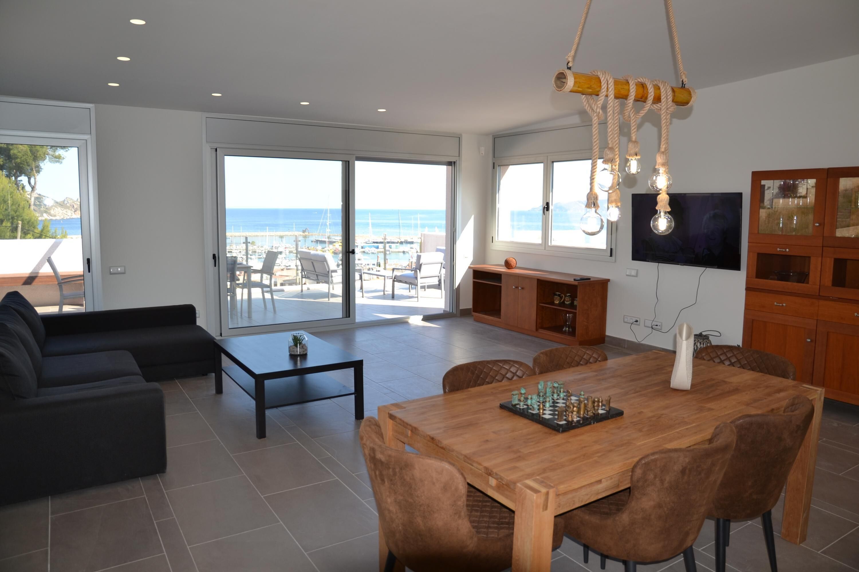 A spacious living area with a wooden dining table, comfortable seating, a television, and a view of the sea through large sliding doors.