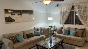 A living room with two beige sofas, a glass coffee table, a ceiling fan, a wall-mounted lamp, and a window with curtains. - Your home away from home! (Cafemba)