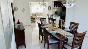 A modern dining area with a long table set for six, surrounded by wooden chairs. A kitchen is visible in the background, and a living area with a sofa and a coffee table is also visible. - Your home away from home! (Cafemba)