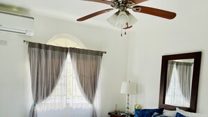 A neatly made bed with white bedding, a wooden headboard, and decorative pillows. A ceiling fan is installed above the bed, and a small bedside table with a lamp is visible. - Your home away from home! (Cafemba)