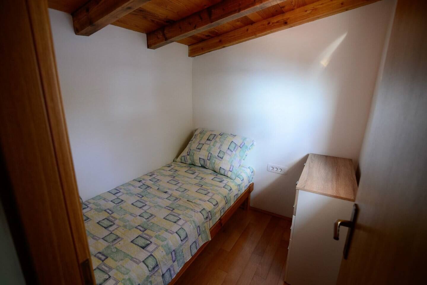 Et lille værelse med en enkelt seng, træloft, et sengebord og en stikkontakt.