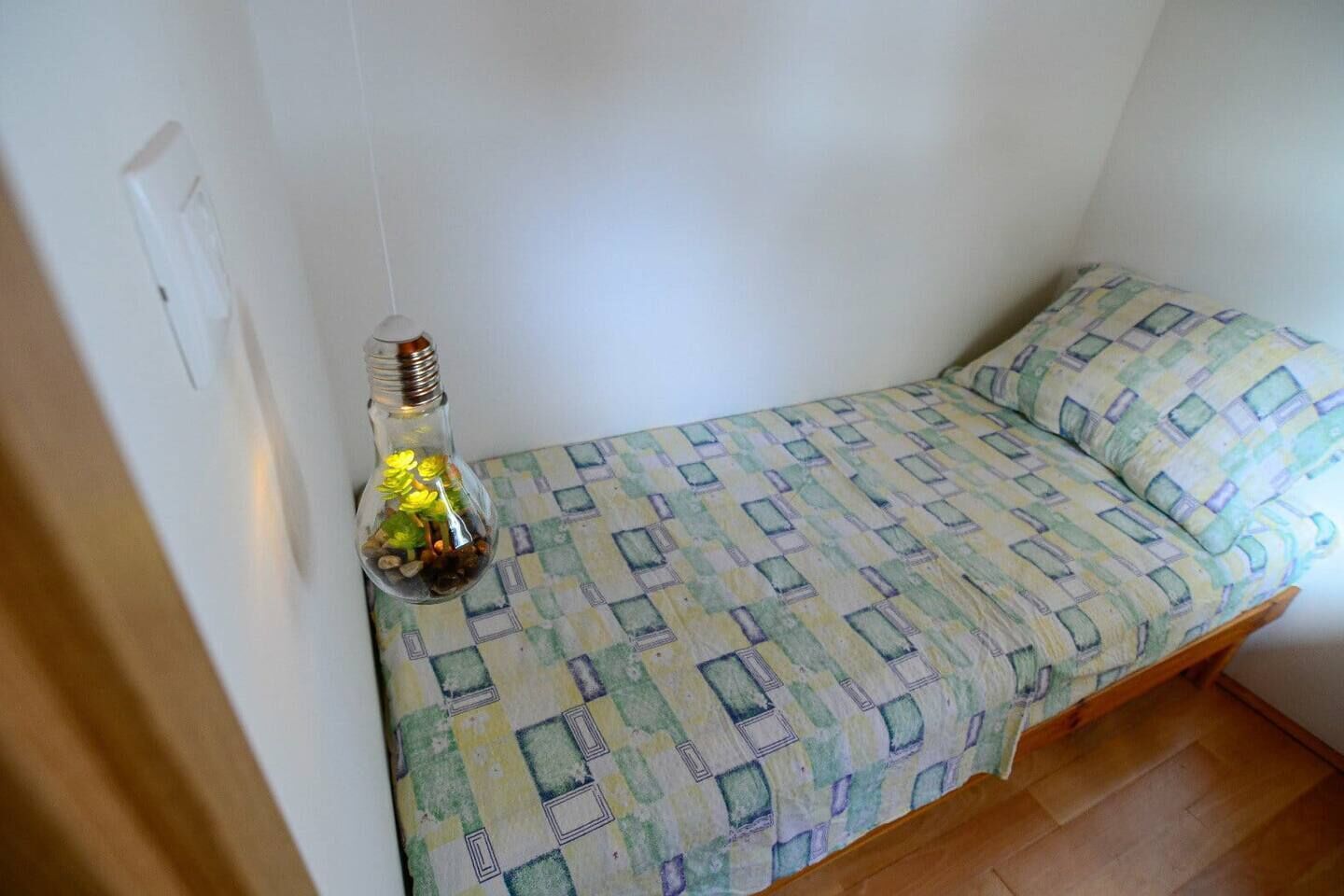 Ein kleines Zimmer mit einem Einzelbett, einer gesteppten Bettdecke, einer Wandleuchte und einem Holzboden.