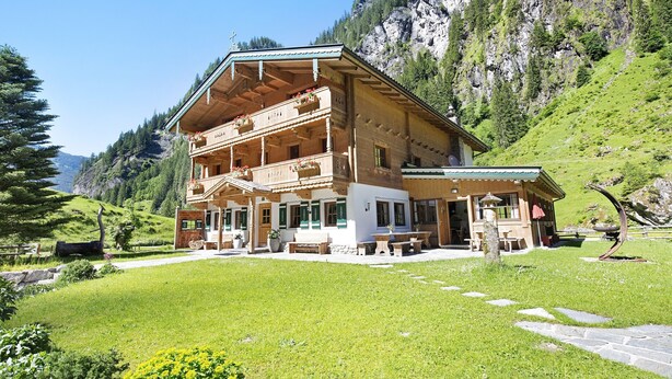 Un chalet de madera con balcón, rodeado de un césped cuidado y con montañas al fondo.