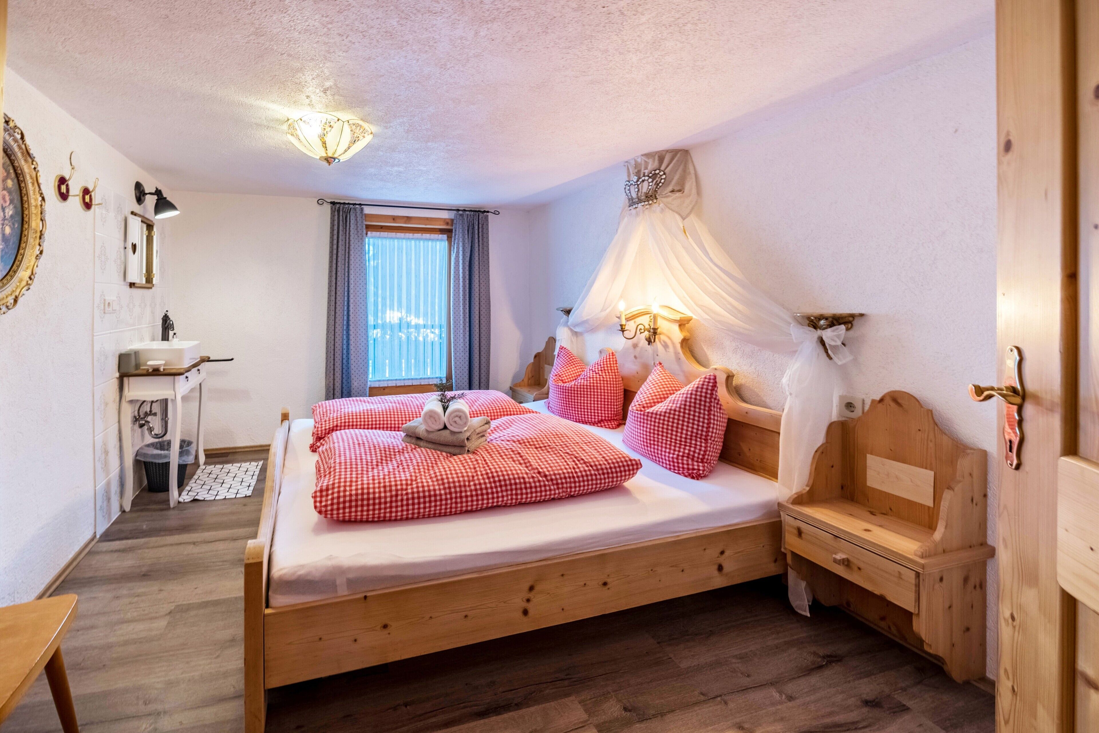 Un dormitorio con cama de madera, una mesita de noche, un pequeño armario de madera, una ventana con cortinas y una lámpara de pared.