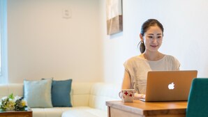 A person sitting at a wooden table with a laptop and a mug, in a room with a white couch and a small table with flowers.