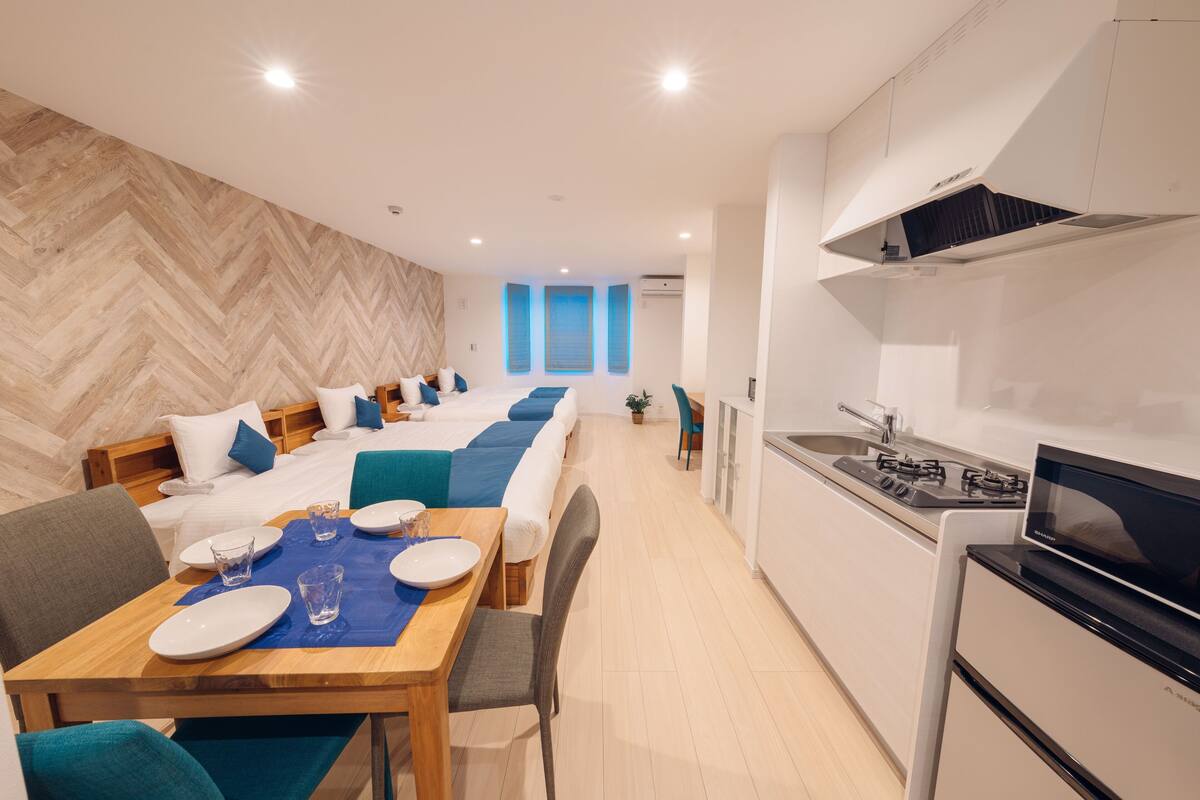 A modern hotel room with a long bed, a dining area with a table and chairs, a kitchenette with a stove and microwave, and a herringbone-patterned wooden floor.