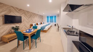 A modern hotel room with a dining area, a bed with blue bedding, a TV mounted on the wall, and a kitchen with a stove and microwave.