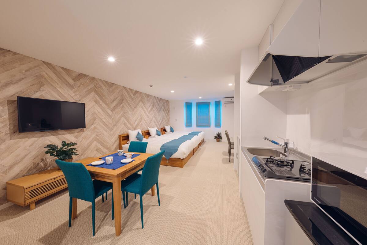A modern hotel room with a dining area, a bed with blue bedding, a TV mounted on the wall, and a kitchen with a stove and microwave.