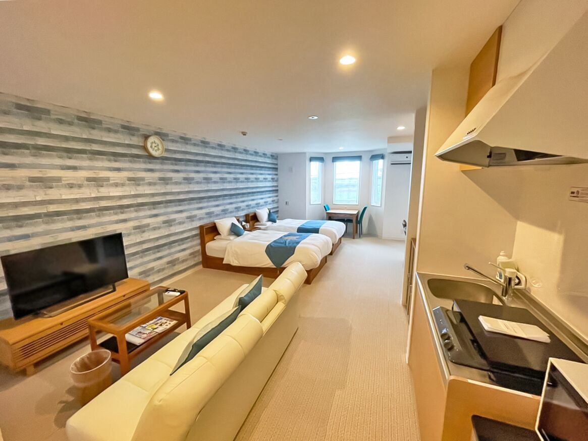 Moderni hotellihuone, jossa on sohva, kaksi sänkyä, televisio, pieni pöytä ja minikeittiö.