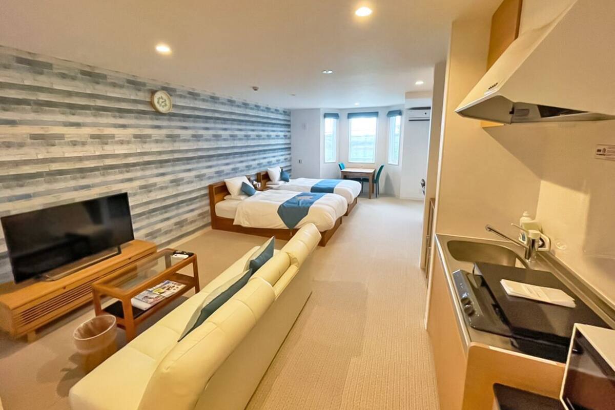 A modern hotel room with a sofa, two beds, a TV, a small table, and a kitchenette.