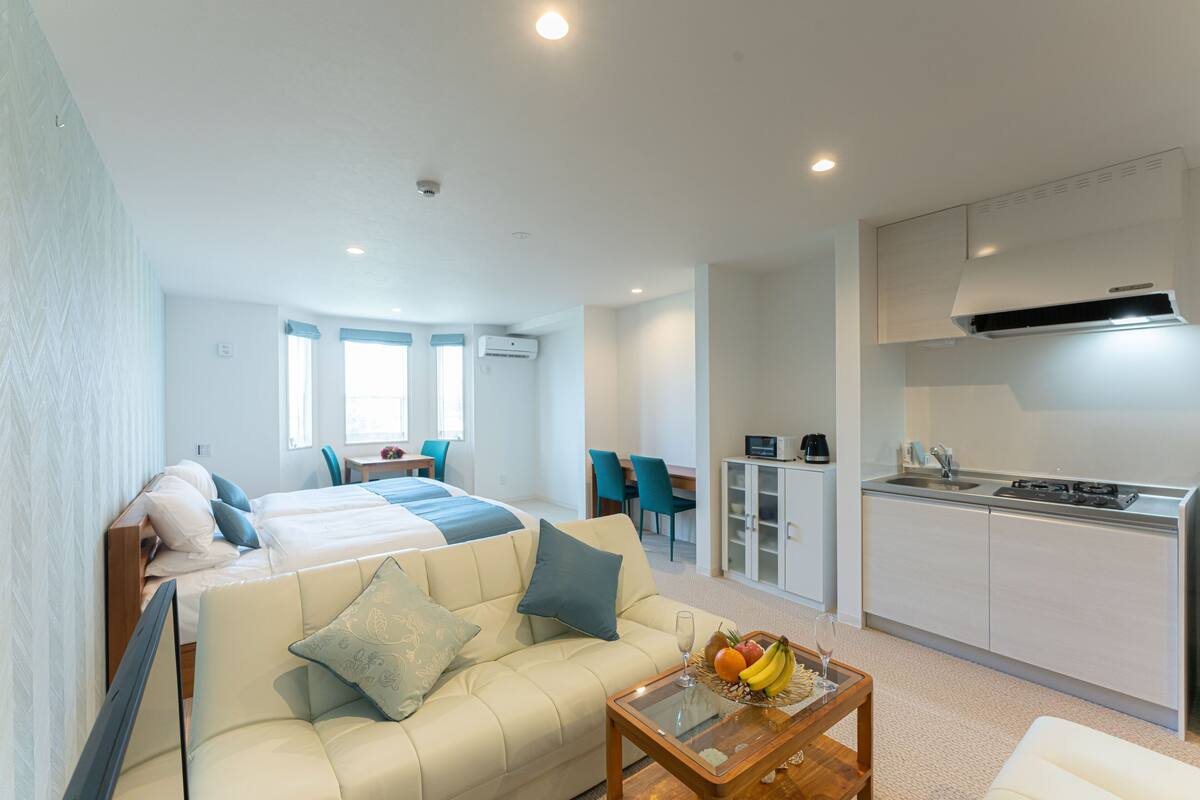 A modern hotel room with a bed, sofa, dining table, and kitchenette.