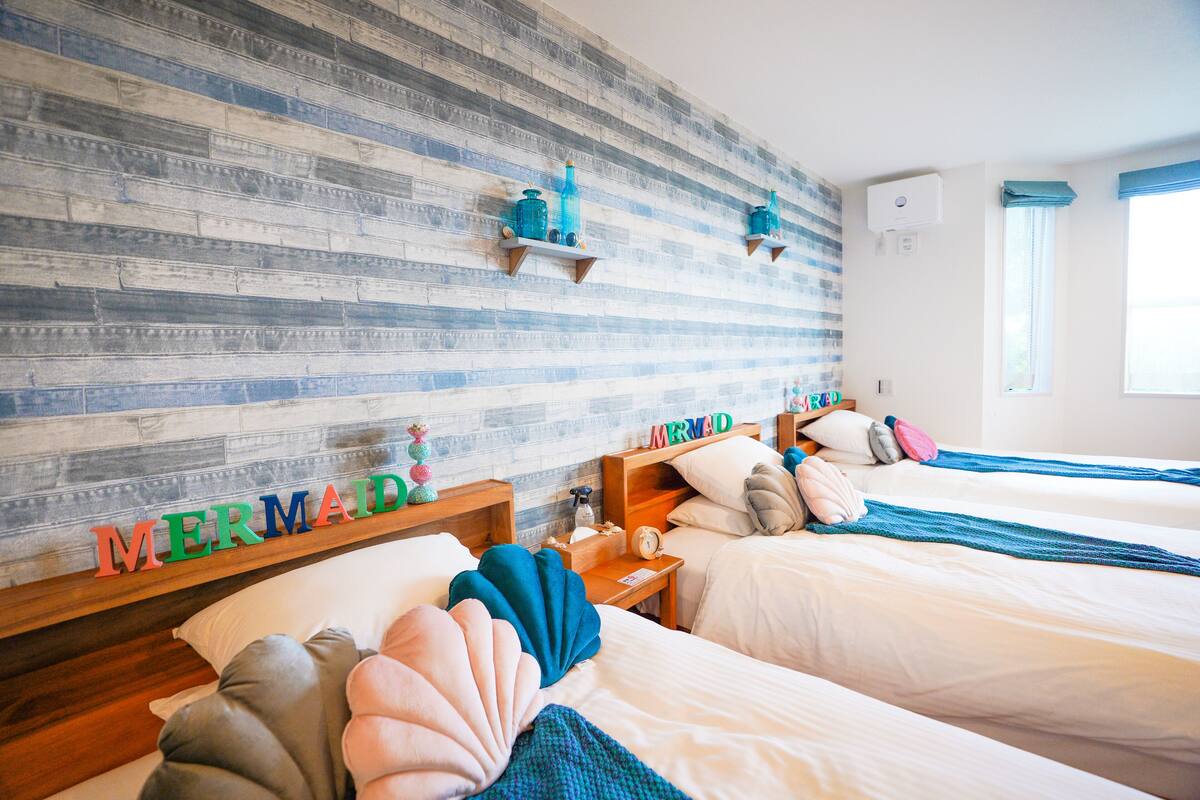 A hotel room with two beds, a wooden wall with a 'MERMAID' sign, and a window with blue curtains.