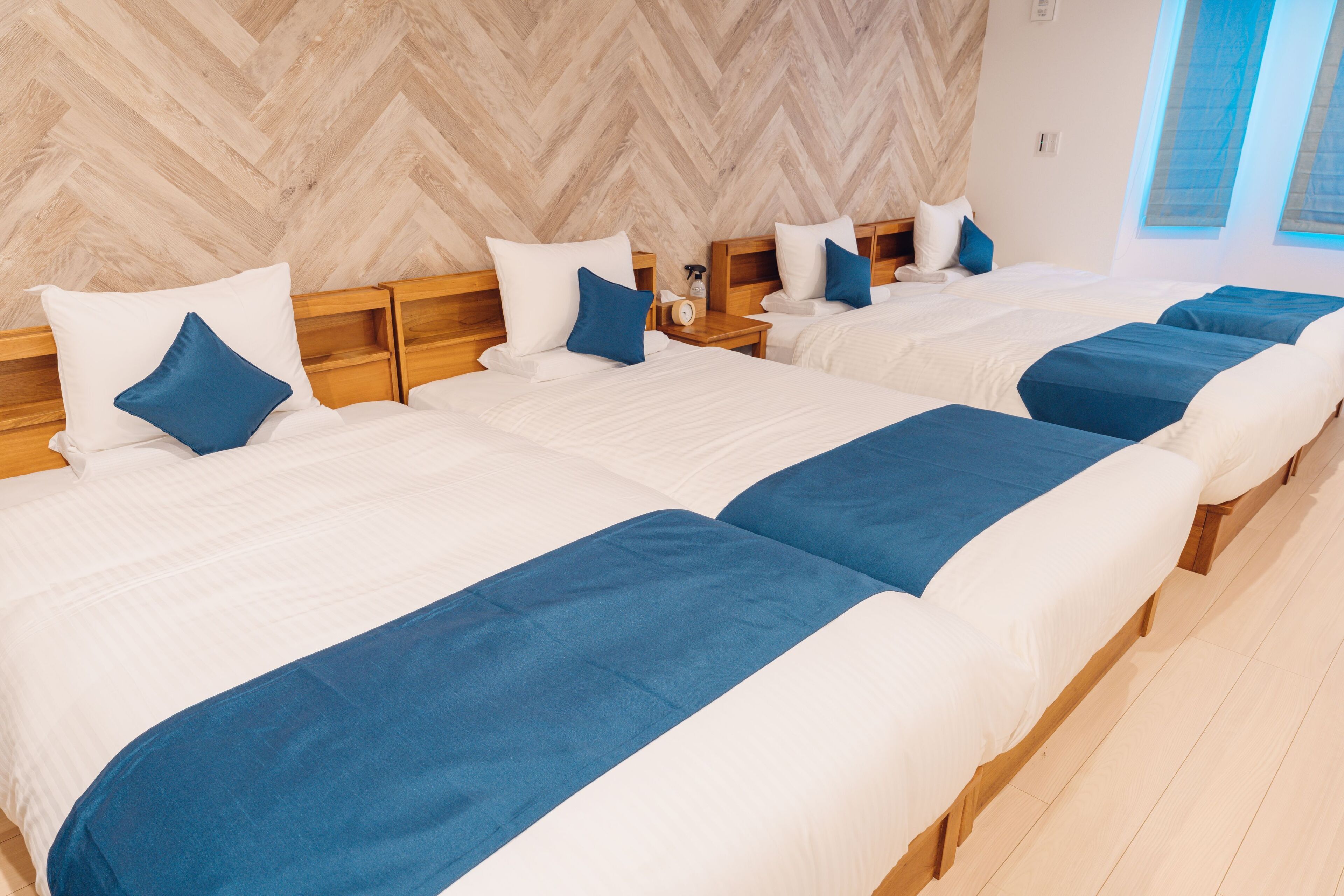 A hotel room with four beds arranged in a row, each with a wooden headboard and white bedding with blue accents.