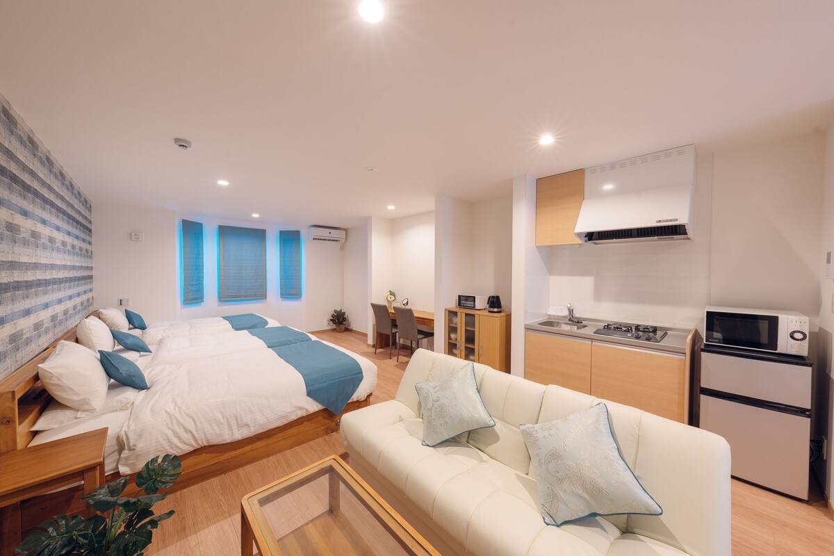 A modern, well-lit room with a bed, a sofa, a small kitchen area, and a dining table.