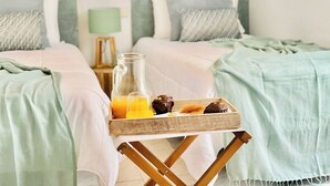 A hotel room with two beds, each with a green headboard and white bedding. A wooden tray with a glass of orange juice, a cookie, and a mug is placed on the floor between the beds.