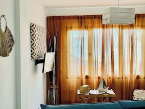 Una habitación con un sofá azul, una mesita de madera con una lámpara, un adorno en la pared y un amplio ventanal con cortinas.