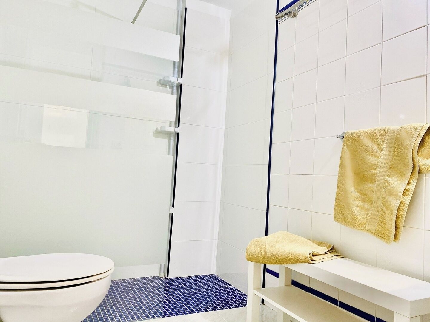 Un baño con inodoro blanco, cabina de ducha de vidrio y un perchero con toalla amarilla.