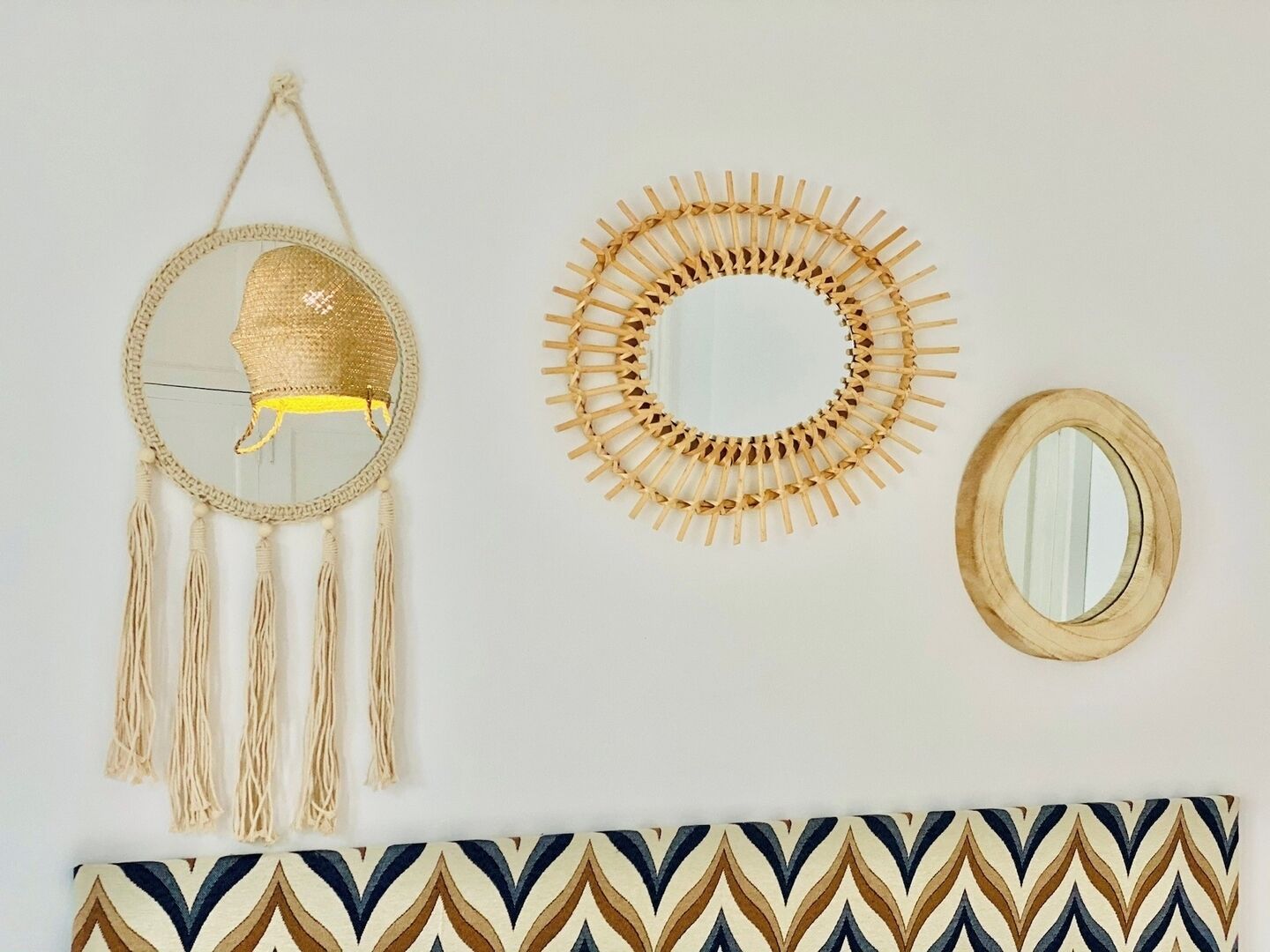 Una pared decorada con tres elementos: un tapiz tejido con flecos, un espejo circular tejido y un pequeño espejo ovalado.