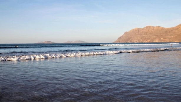 A coastal beach scene with waves, a clear sky, and distant mountains.
