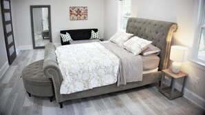 Un dormitorio con una cama grande, un reposapiés, una mesita de noche con una lámpara, un espejo y una ventana.