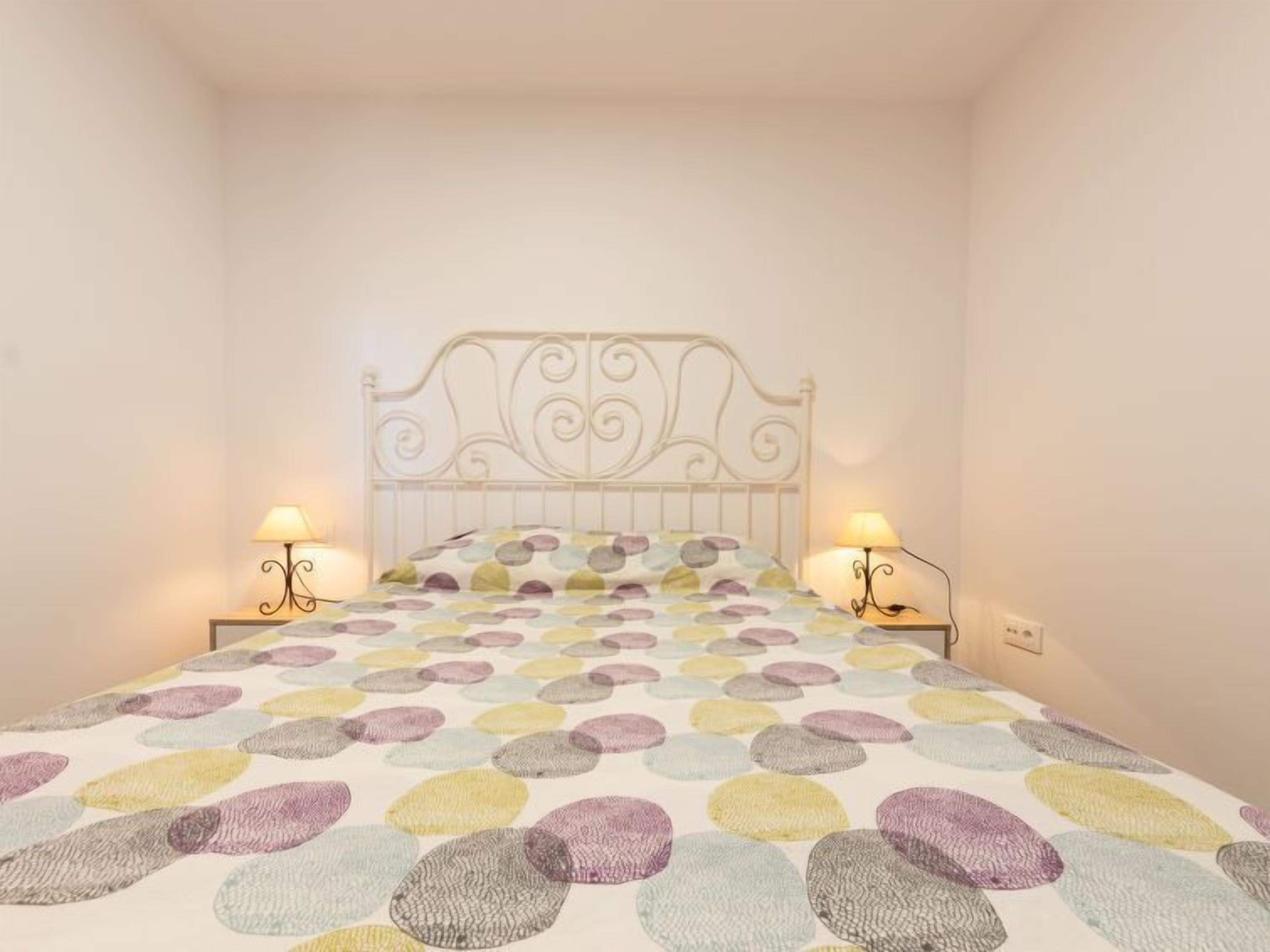 Un dormitorio con estructura de cama metálica blanca, colcha de cama de patrón circular y colorido, y dos lámparas de noche.