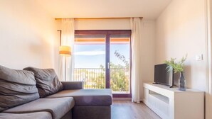 A modern living room with a grey sofa, a flat-screen TV on a white cabinet, and a balcony with a view of greenery. - Marina Pándols by Interhome (Torredembarra)