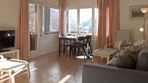 En hyggelig stue med TV, sofa, spisebord med stole og store vinduer med gardiner.