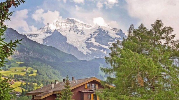 En toetages chalet med et skråt tag, omgivet af frodig vegetation og sneklædte bjerge i baggrunden.