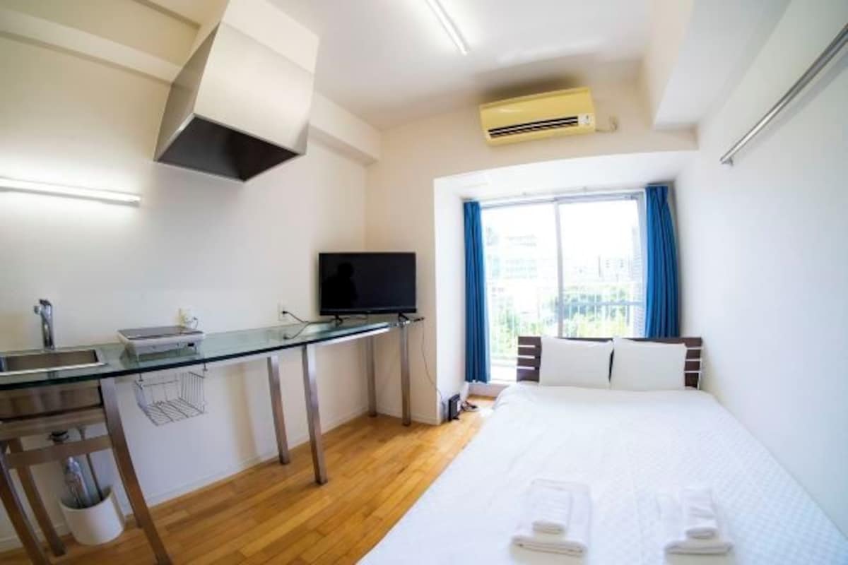 A hotel room with a bed, a desk with a TV, a window with curtains, and an air conditioning unit.