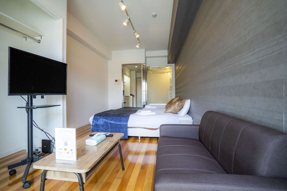 A hotel room with a flat-screen TV, a wooden desk with a sign, a bed with a blue blanket, a gray sofa, and a mirror on the wall.