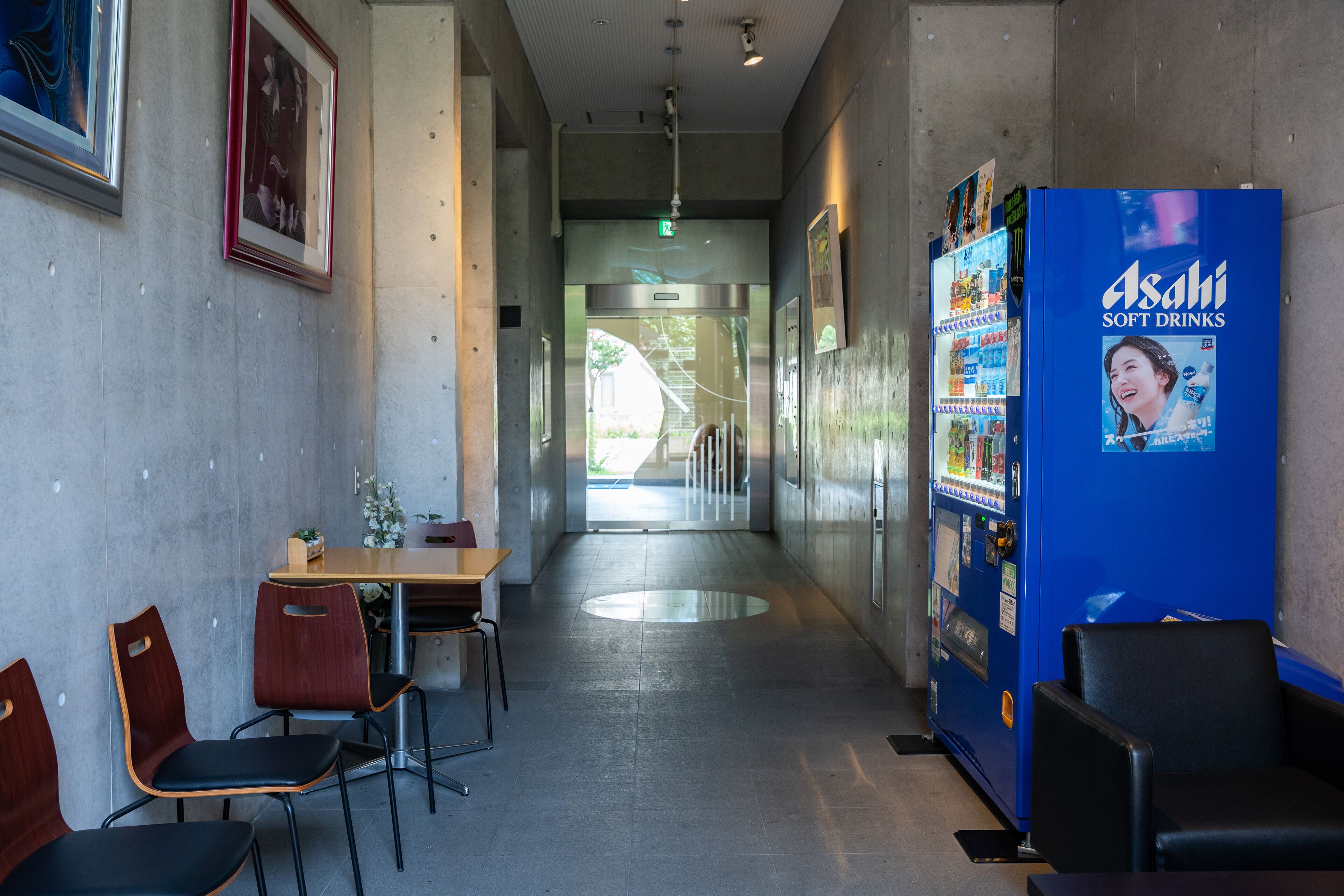 A hallway with a vending machine, chairs, a table, and framed pictures on the walls.