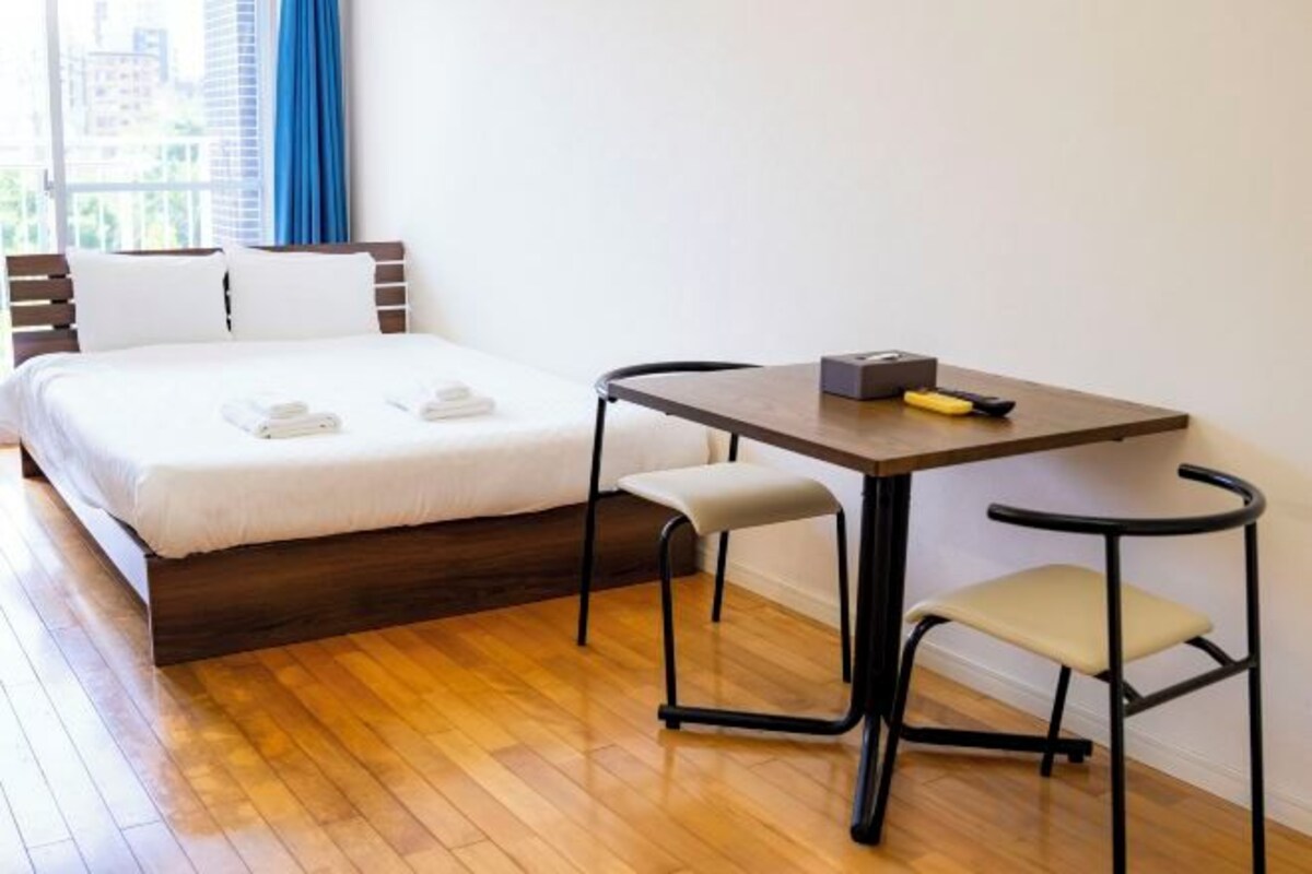 A hotel room with a bed, a table, two chairs, and a window with a view of buildings.