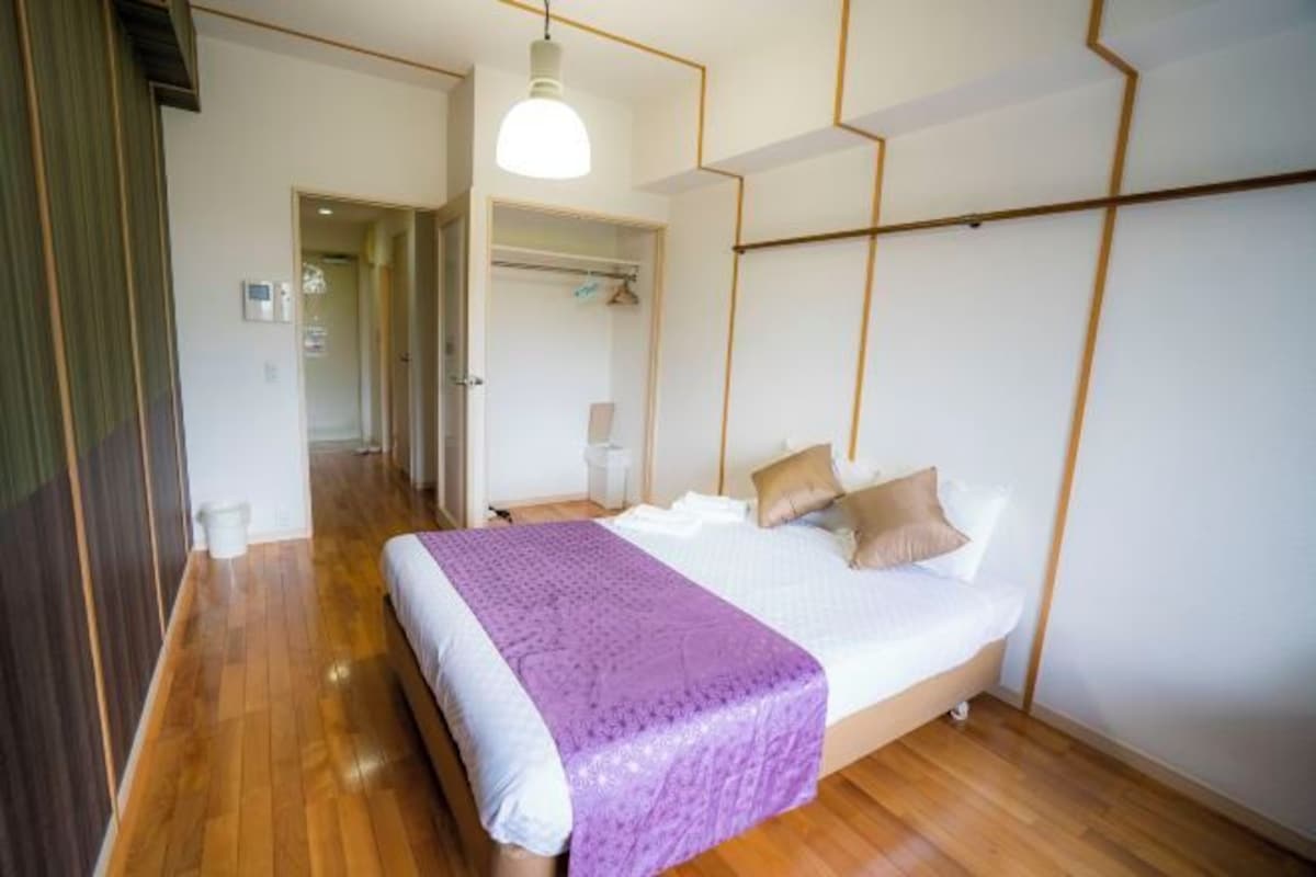 A bedroom with a wooden floor, a bed with a purple blanket, and a white ceiling with geometric lines.