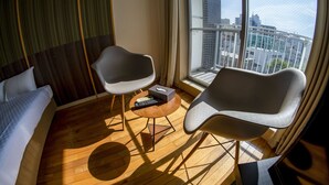 A hotel room with a bed, two chairs, a small table with a remote control, and a large window offering a city view. - S-Presso Namba Parks Street  (Osaka)