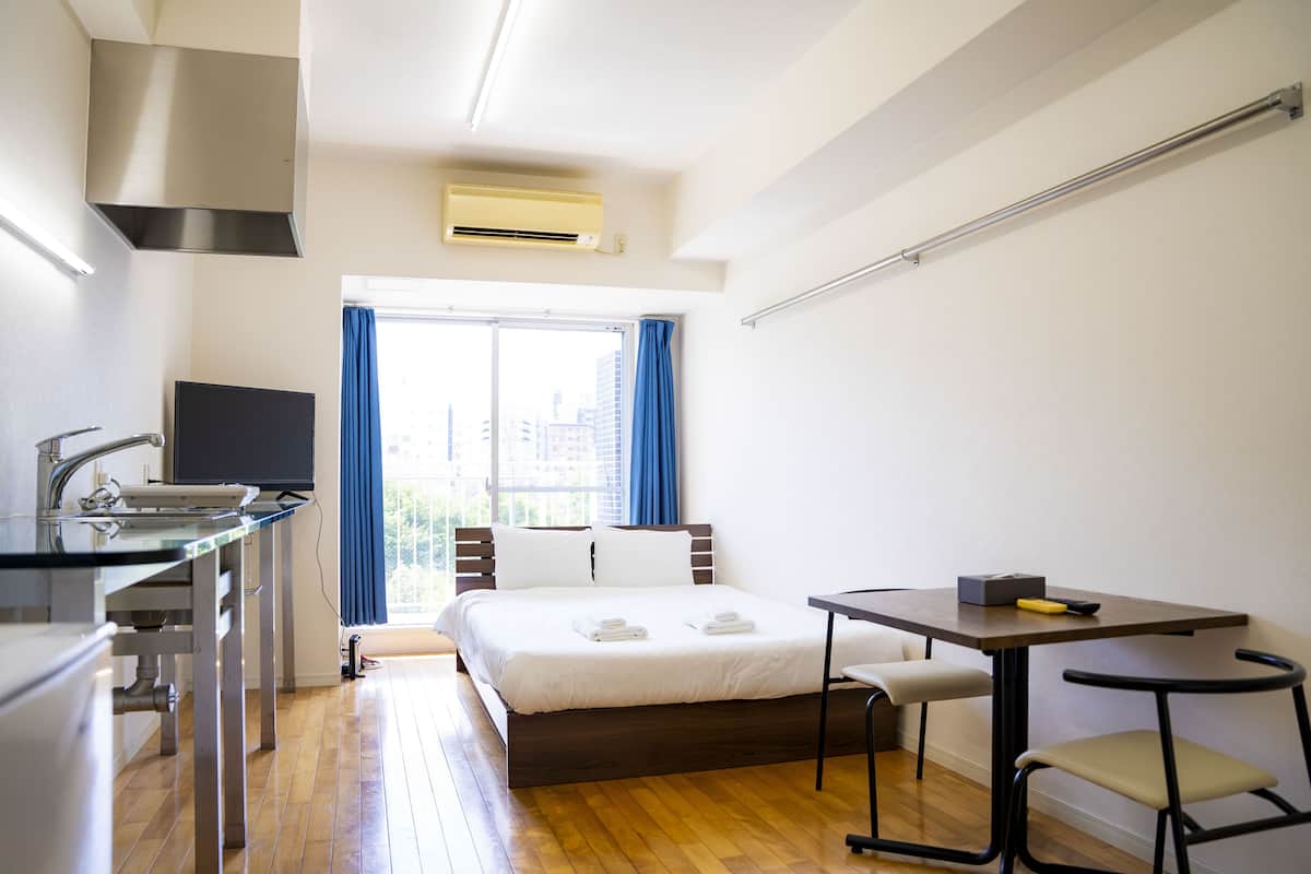 A modern hotel room with a bed, a small desk, a chair, a window with curtains, and a kitchen area with a sink and a television.