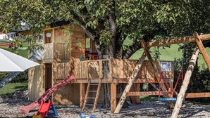 A wooden playhouse with a slide and climbing area, surrounded by trees and a gravel ground. - 3/4/5/7 Gamsbleame for 1-2 pers., 21 m2 (Bischofswiesen)
