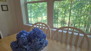 Een eettafel met een vaas blauwe hortensia's, houten stoelen en een raam met uitzicht op bomen.