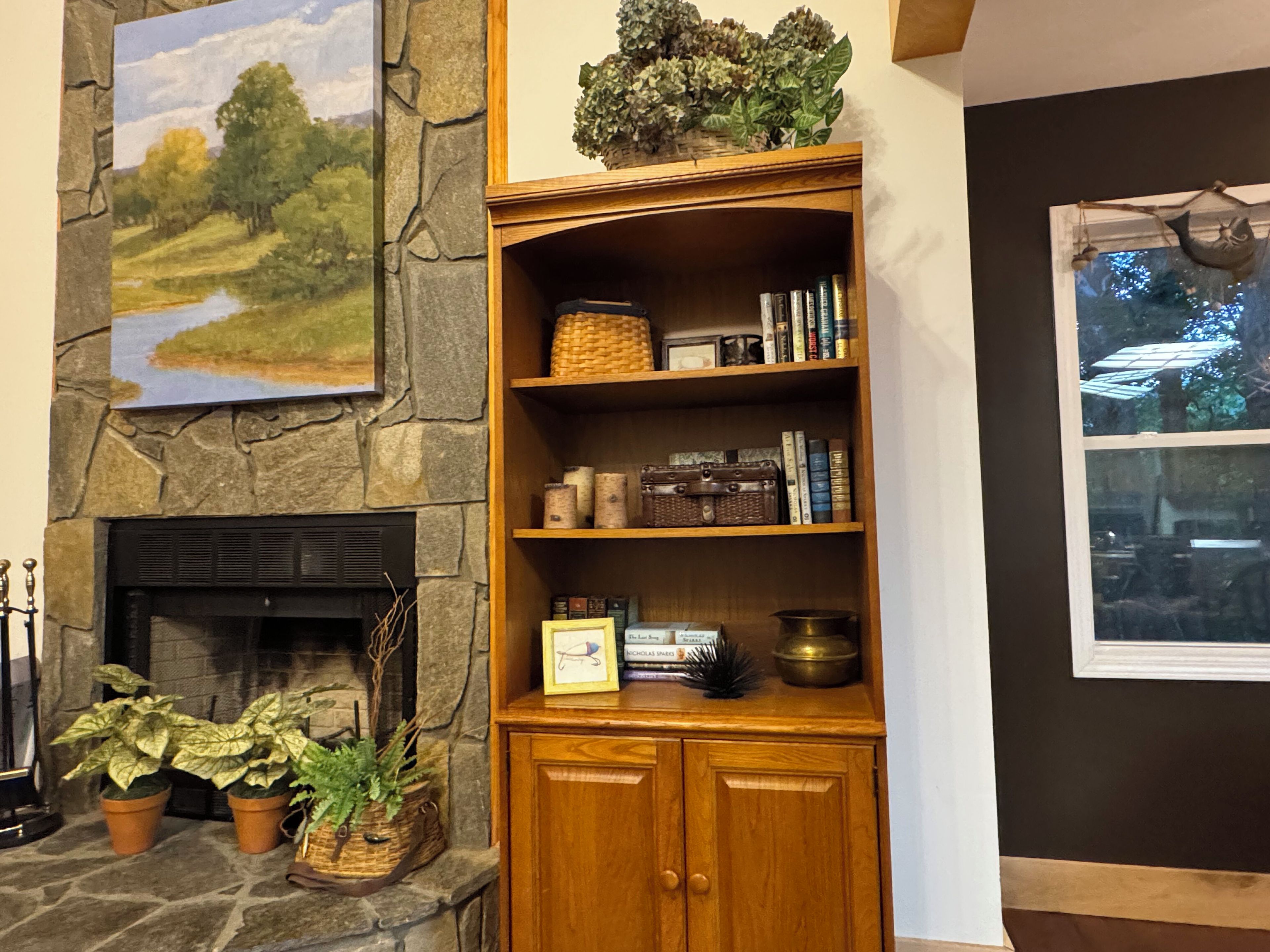 A stone fireplace with a mantel, a wooden bookshelf with decorative items, and a framed landscape painting above it.