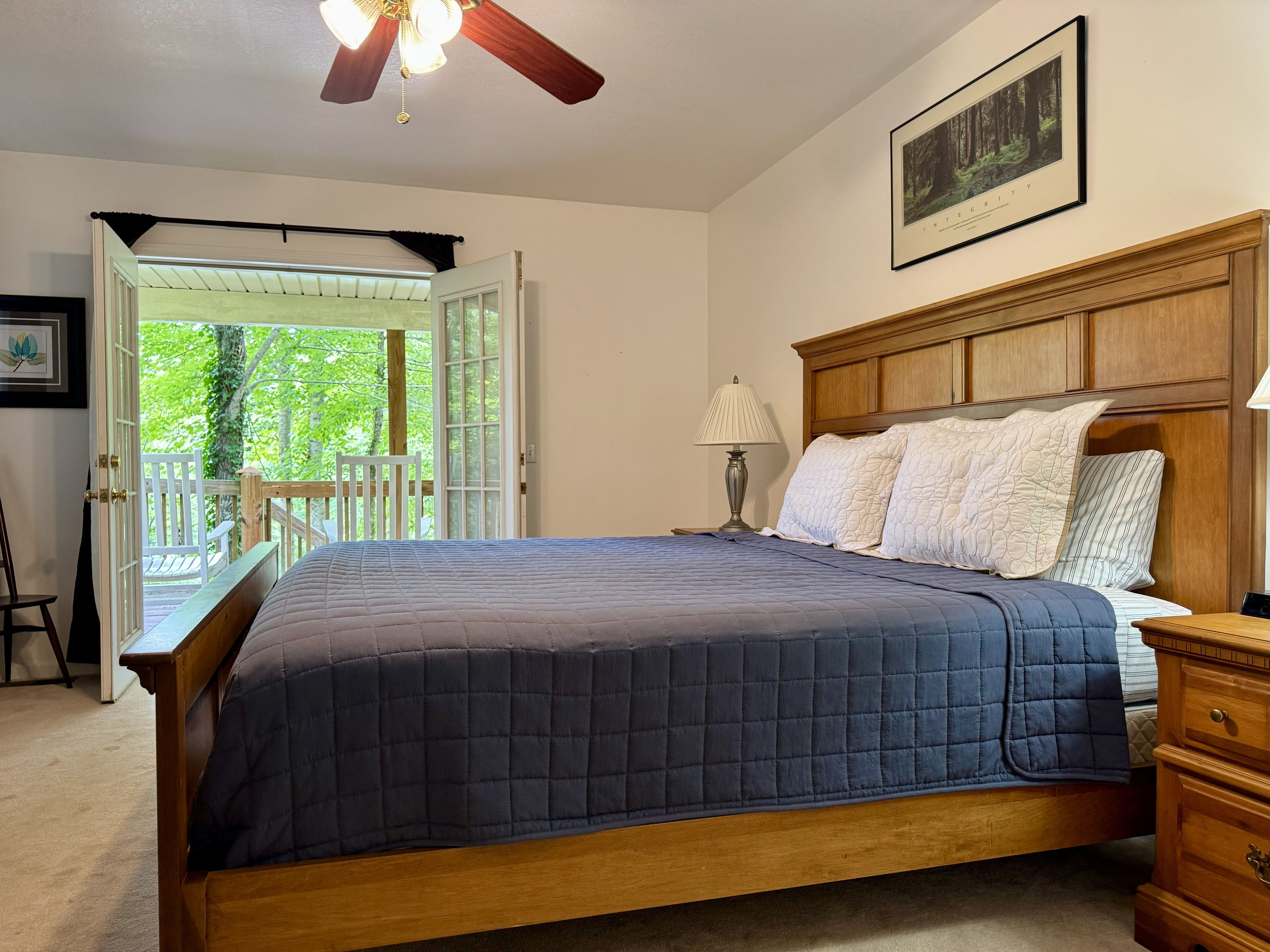 Un dormitorio con una cama grande, una mesita de noche, un cabecero de madera, un ventilador de techo y vistas al exterior.