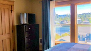 Un dormitorio con cama, cómoda, luz de techo, ventana con cortinas y vistas a un lago y edificios.