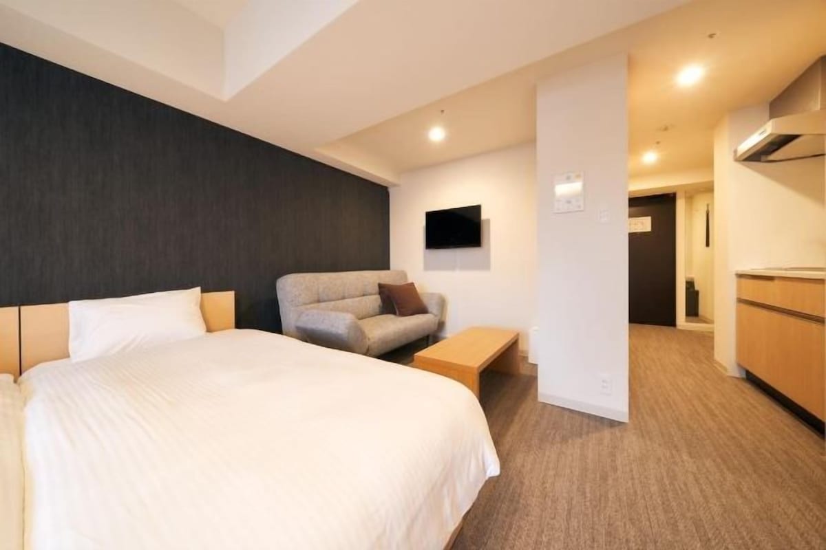 A hotel room with a large bed, a sofa, a small table, and a kitchenette area.