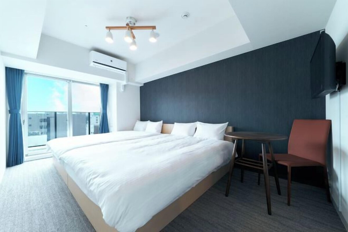 A modern hotel room with a large bed, a small table, and a chair. There is a TV mounted on the wall and an air conditioning unit on the ceiling.
