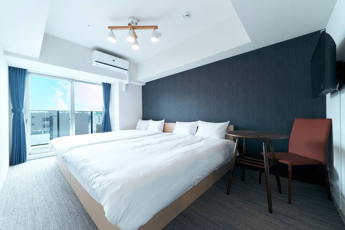 A modern hotel room with a large bed, a small table, a chair, a TV, and a balcony with curtains.