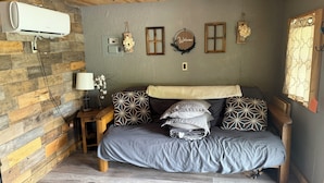 A room with a bed, a wall-mounted air conditioner, a nightstand with a lamp, and a wall with a 'Welcome' sign and decorative items. - Cottonwood Cabin - Cozy Retreat w/ Fire Pit & 150 Acres (Gridley)