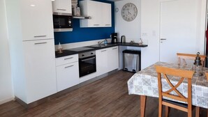 Una cocina compacta con armarios blancos, microondas, estufa y lavamanos. Hay un reloj en la pared y una mesa con sillas en el comedor.