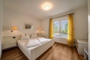 A bedroom with a bed, bedside lamps, a painting, a window with curtains, and a desk.