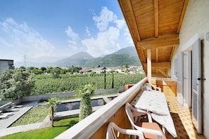 A balcony with a table and chairs, overlooking a garden and a view of mountains and buildings.