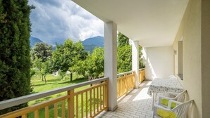 Un balcón con vistas a un paisaje verde y montañas. Hay una mesa con sillas y una lámpara de mesa pequeña.